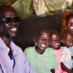Eine Familie lächelt in die Kamera. Ganz vorne sitzt der Vater, David. Er hat eine Sehbehinderung und trägt eine Sonnenbrille. Neben ihm sitzt sein fünfjähriger Sohn Peter. Er hat ein eingeschränktes Sehvermögen und Schwierigkeiten in der Schule. Gemeinsam mit ihrer Familie sind sie aus dem Sudan geflüchtet und leben in Flüchtlingslager Mangatten in Juba im Südsudan.